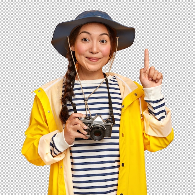Schöne brünette Frau mit asiatischem Aussehen fotografiert während einer Wanderung mit Retro-Kamera trägt einen gestreiften Pullover-Hut und Regenmantel mit dem Zeigefinger über einer gelben Wand