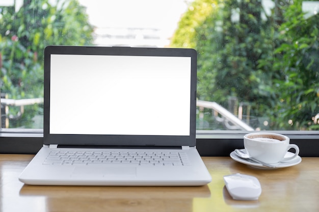 Simulacros de pantalla en blanco de la computadora portátil con una taza de café en la mesa de madera con trazado de recorte fondo borroso.