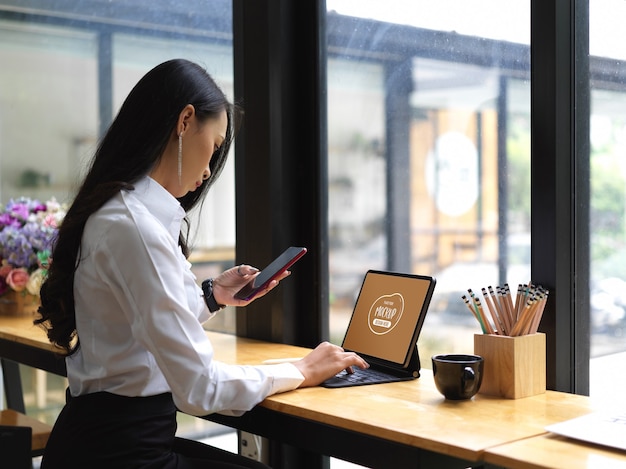 Toma recortada de mujer usando un teléfono inteligente simulado mientras trabaja con una tableta digital simulada en el café