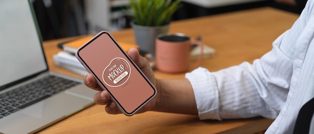 Vista ravvicinata della mano maschio che tiene per mostrare mock up smartphone mentre è seduto al tavolo da lavoro