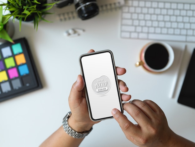 Vista superior de um jovem segurando um smartphone simulado em seu escritório com suprimentos de escritório