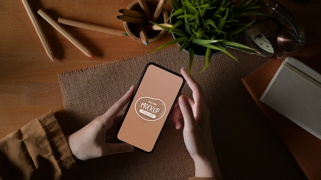 Vue aérienne de la femme à l'aide d'une maquette de smartphone sur une table d'étude avec de la papeterie et un pot de fleurs