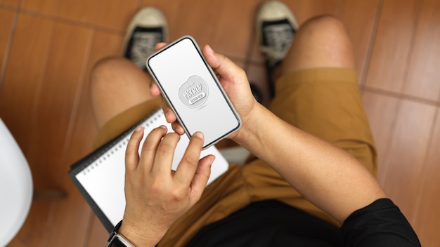 Vue aérienne d'un jeune homme travaillant sur son idée tout en touchant une maquette de smartphone dans sa chambre