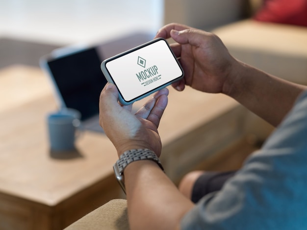 Vue latérale des mains mâles tenant une maquette de smartphone horizontal tout en vous relaxant dans le salon