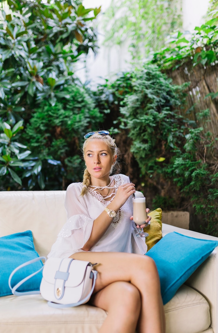 Ücretsiz Fotoğraf stylish blonde young woman sitting on sofa with crossed legs holding glass of latte macchiato