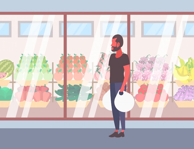 Hombre casual con bolsas de compras caminando delante de frutas y verduras orgánicas frescas supermercado moderno escaparate de cristal chico cliente de longitud completa personaje de dibujos animados plana