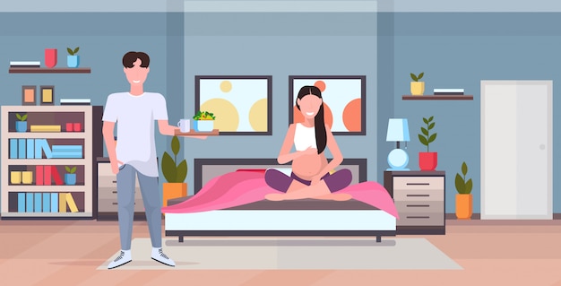 Hombre trayendo la bandeja de desayuno con comida para la mujer embarazada sentada en la cama moderna casa dormitorio interior futuros padres enamorados feliz concepto de familia horizontal de longitud completa