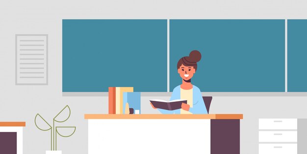 Profesor sonriente sentado en el escritorio mujer comprobando el cuaderno de los estudiantes frente a la pizarra verde concepto de educación aula moderna retrato interior horizontal