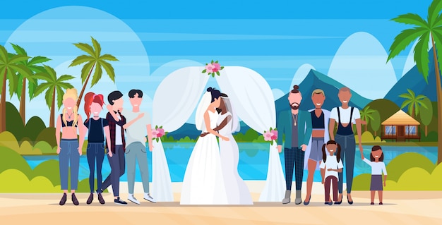 Casal recém-casados lésbicas em vestido branco em pé atrás de arco mesmo gênero casamento homossexual família celebrando conceito tropical ilha seascape paisagem fundo comprimento total horizontal