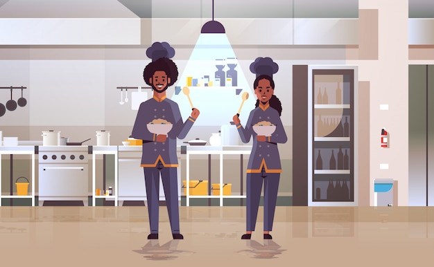Vetor cozinheiros casal chefs profissionais segurando placas com mingau trabalhadores afro-americanos em pratos de degustação uniforme, cozinhando o conceito de comida moderno restaurante cozinha interior