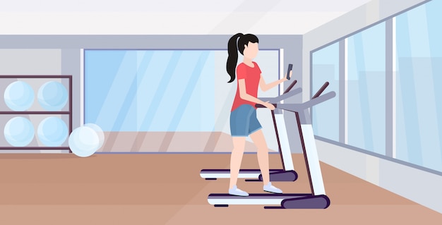 donna che funziona sulla ragazza della pedana mobile che per mezzo dello smartphone mentre preparando orizzontale integrale interno dello studio moderno della palestra di concetto digitale di dipendenza dell'aggeggio di allenamento