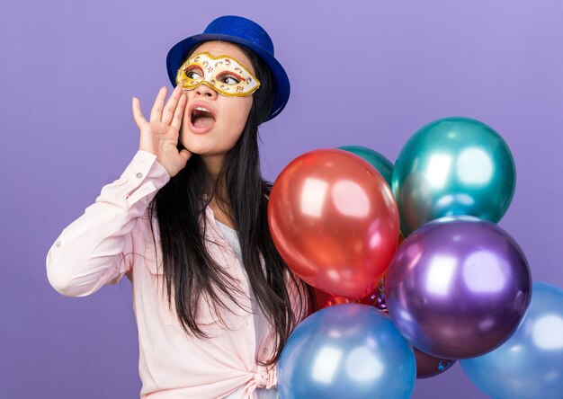 Aan de kant kijkende jonge mooie vrouw met een feesthoed en een maskerade-oogmasker met ballonnen die iemand belt die op een blauwe muur is geïsoleerd