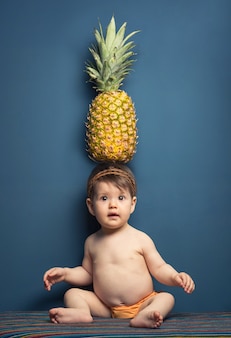 Aanbiddelijk gelukkig babymeisje dat een ananas boven haar hoofd op een blauwe achtergrond houdt