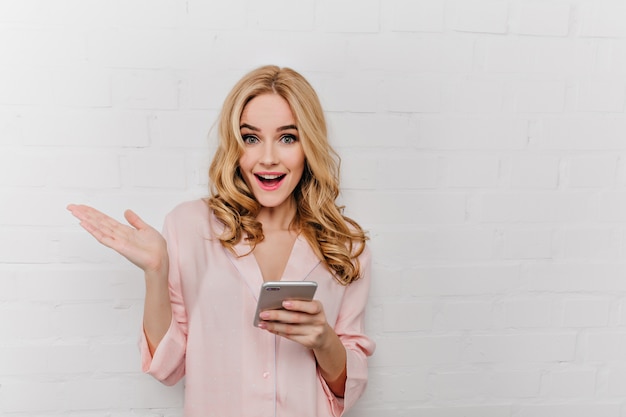 Aanbiddelijk meisje dat met in hand telefoon positieve emoties uitdrukt. Aantrekkelijke blonde vrouw in katoenen roze nachtkleding met smartphone in de buurt van dichtgemetselde muur.