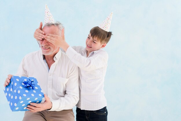 Aanbiddelijke glimlachende jongen die verraste gift geeft aan zijn grootvader door zijn ogen tegen blauwe achtergrond te behandelen