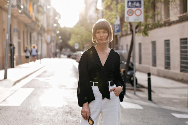 Aanbiddelijke mooie Europese vrouw met kort kapsel die vrijetijdskleding draagt die voor camera in zonneschijn op de zomerstraat in warme zonnige dag stelt