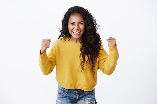 Aangemoedigde en gemotiveerde schattige vrouw in gele trui die handen opsteekt, vuistpomp van geluk, glimlachend goed nieuws horen, overwinning vieren, enorme weddenschap winnen, witte muur