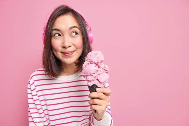 aangenaam ogende jonge Aziatische vrouw met donker haar houdt groot aardbeienijs en kijkt weg en draagt bedachtzaam stereohoofdtelefoons geïsoleerd over roze muurkopieruimte.