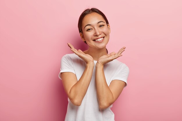 Aangenaam uitziend Aziatisch vrouwelijk model glimlacht blij, spreidt de handpalmen in de buurt van het gezicht, drukt positieve emoties uit, draagt een wit t-shirt, heeft een aantrekkelijk uiterlijk, een gezonde huid, geïsoleerd op een roze muur