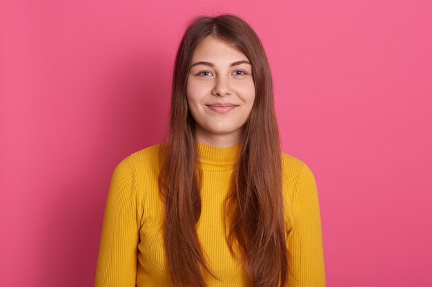 Aangenaam uitziende blanke vrouw met lang haar, gekleed in een gele casual shirt, met een goed humeur, kijkend naar de camera