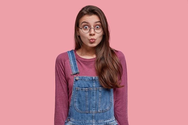 Aangenaam uitziende brunette vrouw pruilt lippen, maakt grimas, draagt paarse trui en denim overall