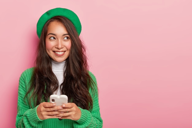 Aangenaam uitziende Koreaanse vrouw heeft een bedachtzame opgewekte uitdrukking, draagt een groene trui en hoofddeksel, poseert met een mobiele telefoon