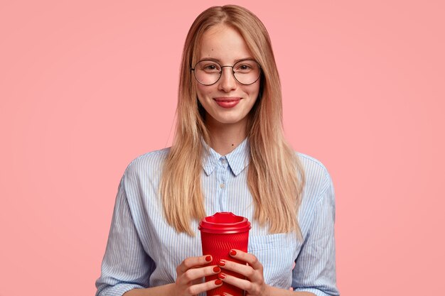 Aangenaam uitziende studente heeft koffiepauze, houdt afhaalmaaltijden rode beker vast, draagt een ronde bril en shirt