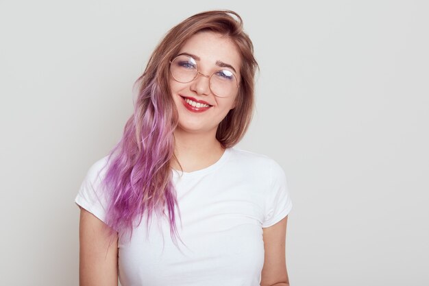 Aangenaam uitziende vrouw met een bril en een wit casual T-shirt die direct naar de camera lacht, heeft lila haar, dat positieve emoties uitdrukt, geïsoleerd over een grijze achtergrond.