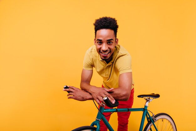 Aangename Afrikaanse man met krullend haar naast de fiets. Binnen schot van extatische zwarte man poseren