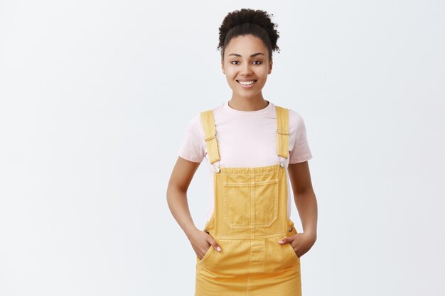 Aangename schattige, donkere vrouwelijke medewerker die klanten helpt bij het vinden van het juiste artikel om te kopen. Vrolijk vriendelijk ogend meisje in trendy gele overall, hand in hand in de zakken en lachend