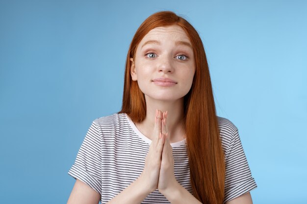 Aanhankelijk schattig aardig roodharig europees meisje dat gunst vraagt beker handen samen bidden gebaar zeg alsjeblieft anticiperen op akkoord of goedkeuring smeken om hulp staande blauwe achtergrond. Ruimte kopiëren