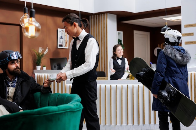 Aankomst van een vrouwelijke toerist met een snowboard in de lobby van het hotel voor het inchecken bij het personeel Jonge ober serveert koffie aan een man op de bank Wintervakantie sfeer met ski-uitrusting Ski resort zorgt voor een gelukkig verblijf