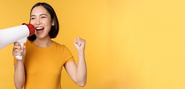 Aankondiging Gelukkige aziatische vrouw die luid schreeuwt tegen megafoon die protesteert met spreker in handen die over gele achtergrond staat