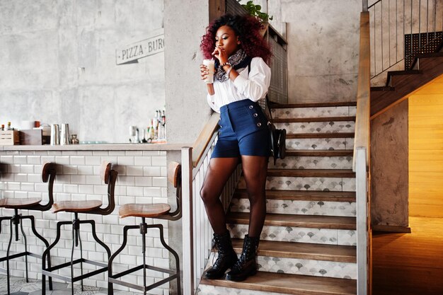 Aantrekkelijk Afrikaans-Amerikaans krullend meisje in witte blouse en blauwe korte broek geposeerd in café met latte bij de hand