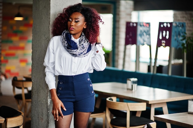 Aantrekkelijk Afrikaans-Amerikaans krullend meisje in witte blouse en blauwe korte broek geposeerd in café