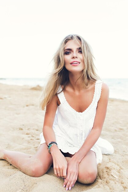 Aantrekkelijk blond meisje met lang haar zit op zand op het strand. Ze kijkt naar de camera.