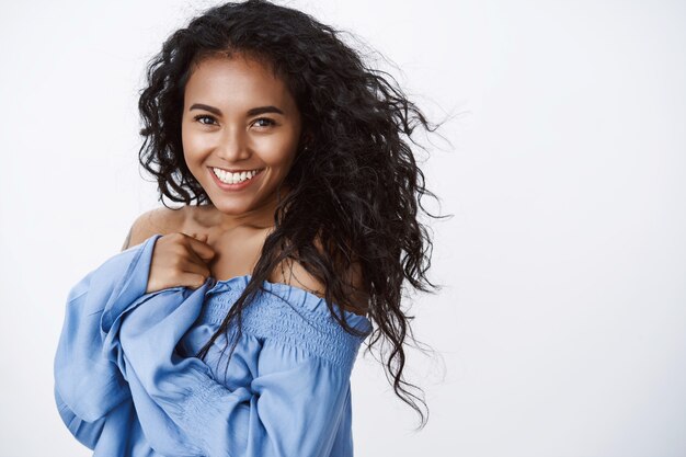 Aantrekkelijk charmant, vrouw met krullend kapsel, blauwe blouse, oprecht lachend en romantisch starende camera, staande witte muur