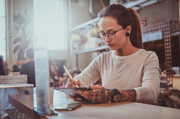 Aantrekkelijk creatief meisje met tatoeages op haar handen zit in café terwijl ze schetst in haar digitale notitieblok.