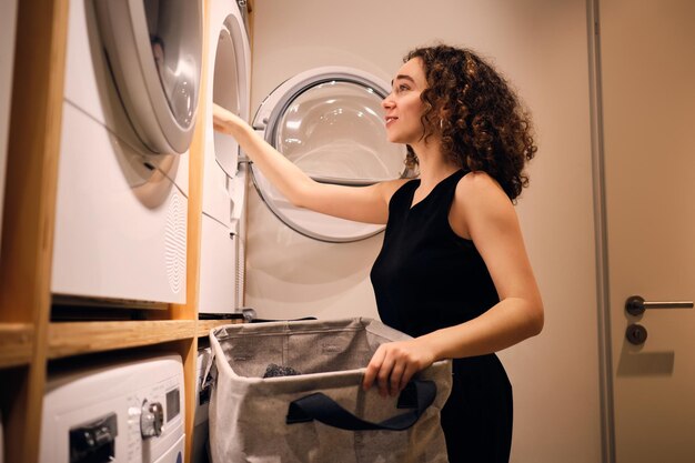 Aantrekkelijk donkerbruin meisje dat dromerig kleren in de wasmachine laadt in moderne zelfbedieningswasserij