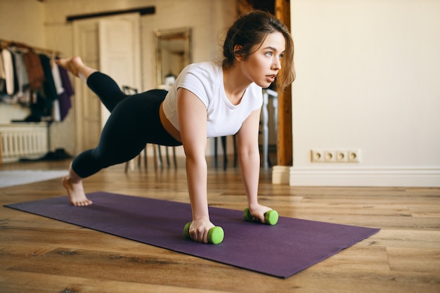 Aantrekkelijk fit meisje doet plank met halters tijdens het trainen op de mat in de woonkamer