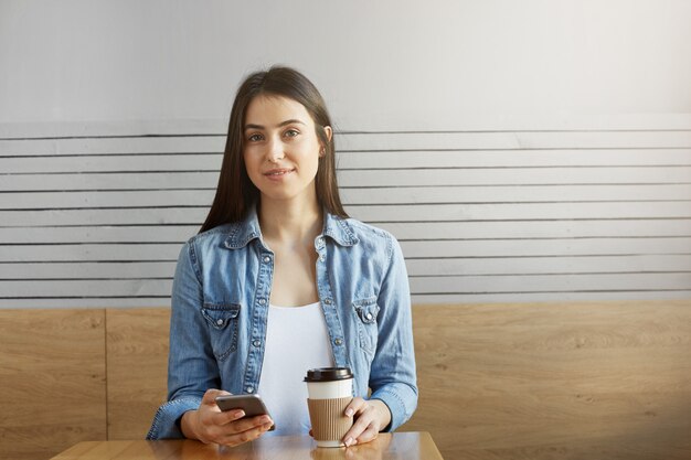 Aantrekkelijk jong meisje met donker haar en modieuze kleren die in koffie zitten, koffie drinken en door foto's van vakantie op haar smartphone kijken.