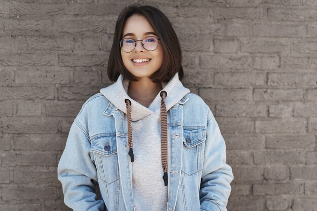 Aantrekkelijk jong meisje met kort haar, bril en street style kleding, leunend op een bakstenen muur.