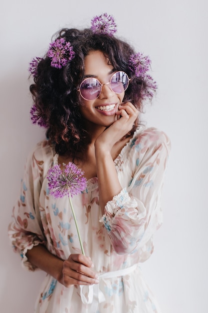 Aantrekkelijk jong vrouwelijk model met bloemen in haar dromerig wegkijken. indoor foto van mooi afrikaans meisje in trendy jurk.