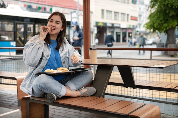 Aantrekkelijk meisje in casual stijl eet een hamburger met koffie zittend op het zomerterras van een café