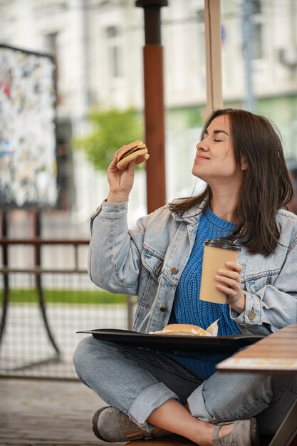 Aantrekkelijk meisje in casual stijl eet een hamburger met koffie zittend op het zomerterras