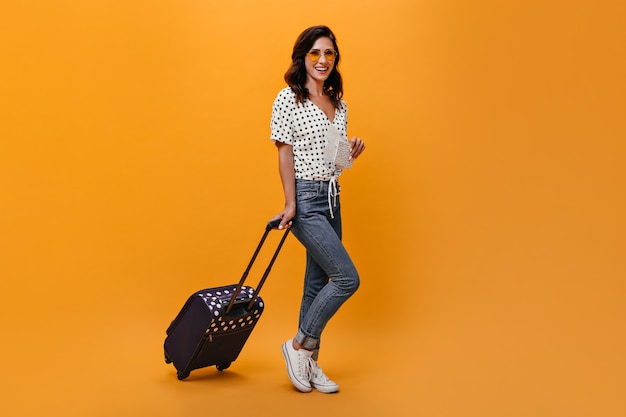 Aantrekkelijk meisje in glazen draagt koffer op oranje achtergrond. Brunette met golvend haar in zonnebril in witte blouse met zwarte polka dots poseren.