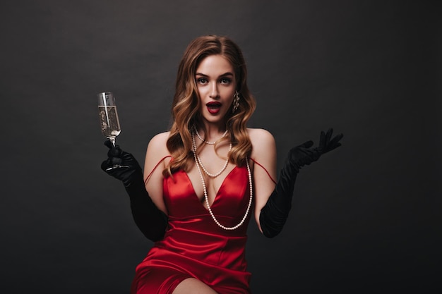 Aantrekkelijk meisje in zijden rode jurk verrast in de camera kijken en champagneglas vasthouden