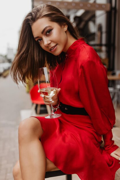 Aantrekkelijk meisje met glanzend steil haar op mysterieuze wijze en verleidelijke looks poseren voor portret Foto van Parijse vrouw in rode jurk met glas witte wijn