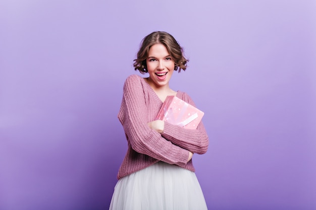 Aantrekkelijk meisje met krullend kapsel nieuw jaar aanwezig houden en glimlachen. elegante vrouw in witte rok poseren met roze geschenkdoos op paarse muur.