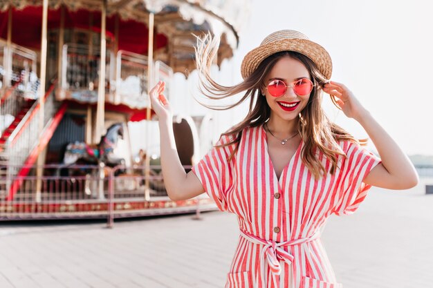 Aantrekkelijk meisje met mooie glimlach gek rond in pretpark. Buiten foto van verfijnde blonde dame in strooien hoed spelen met haar haren naast carrousel.